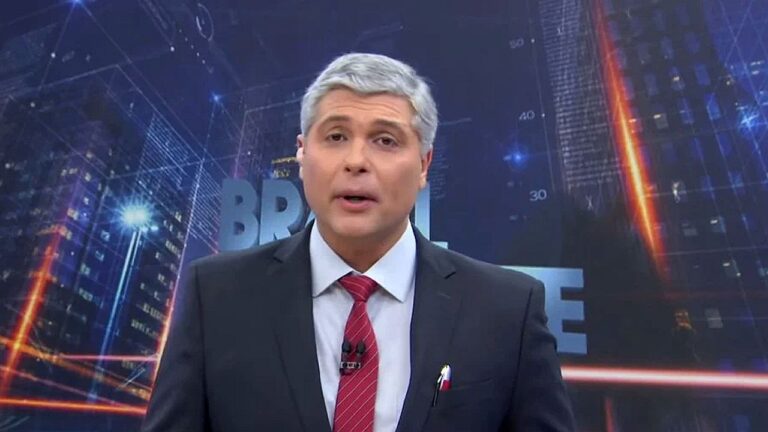 Homem de cabelos grisalhos apresenta um telejornal em estúdio, usando terno escuro e gravata vermelha, diante de telão com gráficos e cenário urbano estilizado ao fundo.