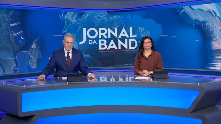 Eduardo Oinegue e Thaís Dias, apresentadores de telejornal, estão sentados atrás de uma bancada iluminada em azul em estúdio moderno. Ao fundo, um grande painel exibe a marca do Jornal da Band sobre um mapa-múndi estilizado. Cada um tem um tablet à frente, e ambos olham para a câmera durante a transmissão.