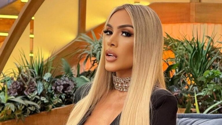 Juju Ferrari com cabelo loiro longo e liso posa com olhar direto usando choker brilhante em ambiente com plantas ao fundo