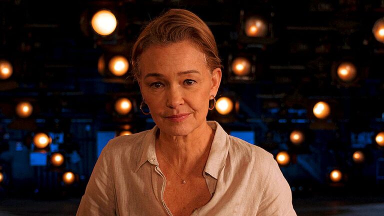 Mulher com expressão séria encara a câmera em estúdio iluminado com luzes ao fundo em cena de série dramática