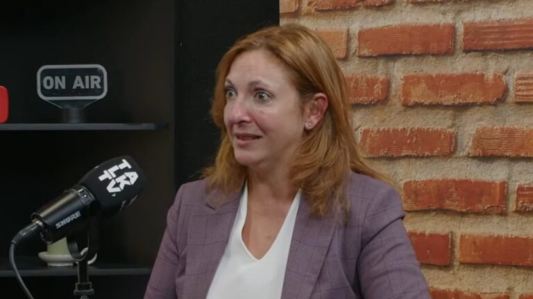 Juliana Algañaraz olha para o lado com expressão atenta, veste blazer xadrez roxo sobre blusa branca e está posicionada à frente de um microfone preto