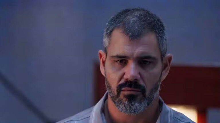 Homem de barba grisalha observa alguém com expressão séria em cena dramática dentro de ambiente interno iluminado com luz suave