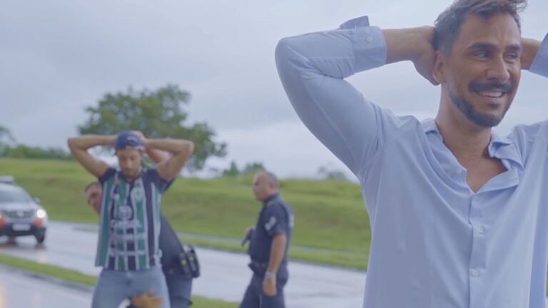 Em primeiro plano, um homem de camisa azul clara está com as mãos atrás da cabeça e sorri enquanto olha para o lado. Ao fundo, em uma estrada ao ar livre, dois policiais parecem revistar ou deter outro homem que também está com as mãos atrás da cabeça. Há um carro de polícia parado na via e uma área gramada com árvores ao redor, sugerindo uma abordagem policial em ambiente externo.
