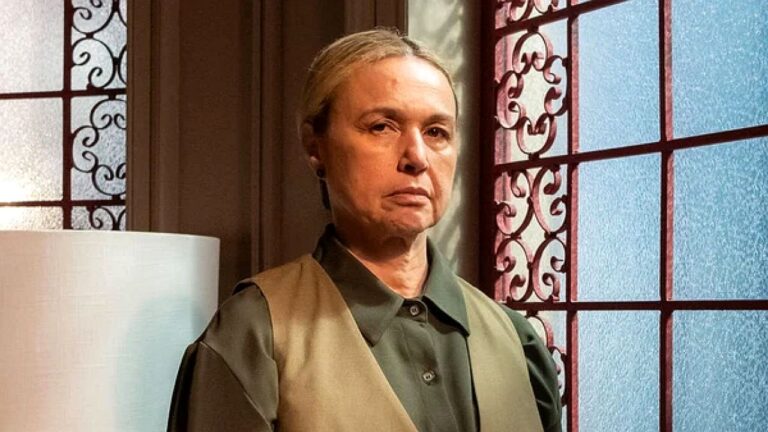 Mulher de aparência séria permanece de pé em ambiente interno ao lado de janela com vitral decorativo e iluminação suave ao redor