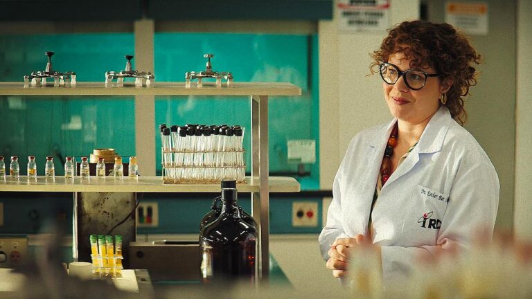 Mulher de jaleco e óculos observa laboratório com tubos de ensaio e frascos em ambiente científico