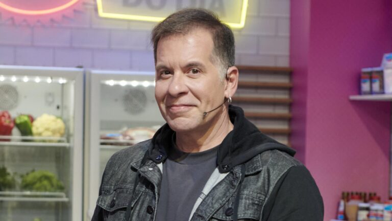 Leandro Hassum sorri para a câmera em estúdio de programa culinário, usando microfone de cabeça e jaqueta escura, com geladeira com alimentos e prateleiras ao fundo.