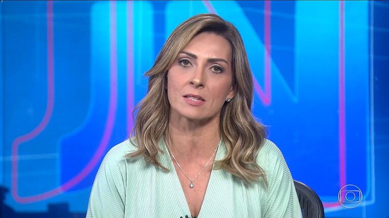 Lídia Pace aparece em um estúdio de televisão, olhando diretamente para a câmera com expressão séria e profissional. Ela tem cabelos médios, levemente ondulados, e usa uma blusa clara com colar discreto. O fundo é azul com elementos gráficos típicos de telejornal, indicando que ela está apresentando ou participando de um programa jornalístico.