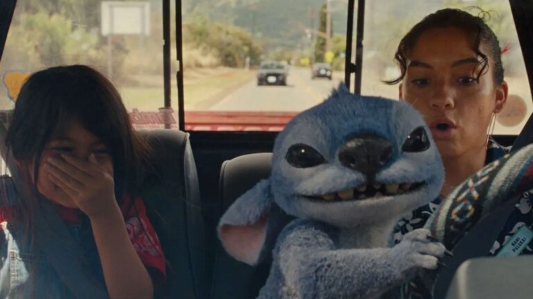 Dentro de um carro em movimento, duas meninas estão sentadas enquanto uma criatura azul animada, com aparência fofa e orelhas grandes, segura o volante e dirige. A jovem à direita parece surpresa, com a boca aberta, enquanto a menina à esquerda cobre o rosto, rindo ou reagindo à situação. Pela janela traseira, é possível ver a estrada e outros carros ao fundo, indicando que o veículo está em trânsito.