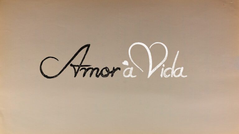Logotipo da novela Amor à Vida com escrita cursiva, palavra Amor em preto e Vida em branco com um coração estilizado no V, sobre fundo bege claro clara