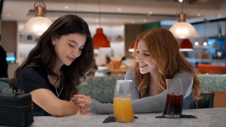 Duas mulheres conversam e sorriem sentadas à mesa de restaurante com copos de suco e refrigerante sobre a mesa