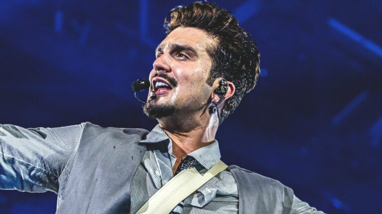 Luan Santana canta durante show usando microfone de headset, cantor veste camisa social cinza, colete, correia de instrumento clara e exibe topete castanho