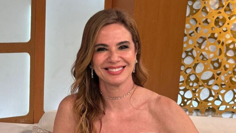 Luciana Gimenez sorri com cabelos castanhos ondulados, apresentadora usa colar choker de brilhantes, brincos pendentes e ombros à mostra em cenário claro