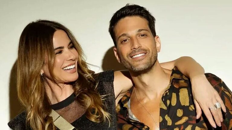 Casal posa sorrindo abraçado para foto em ambiente interno com iluminação suave ao fundo