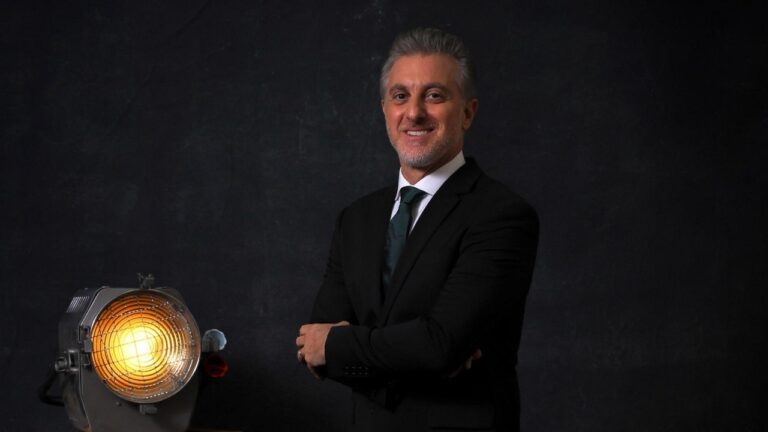 Apresentador Luciano Huck sorri de terno preto e gravata verde escura, posa com braços cruzados ao lado de um refletor de luz aceso em cenário de fundo preto