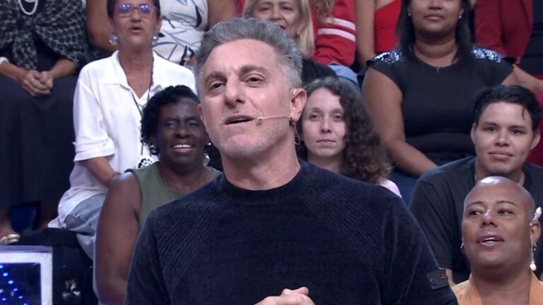 Luciano Huck de suéter preto no palco do Domingão com Huck, sorrindo e falando ao microfone de lapela, com plateia diversificada ao fundo no cenário