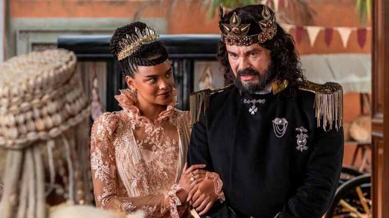 Homem e mulher com coroas e trajes reais conversam em cena de época com figurinos luxuosos