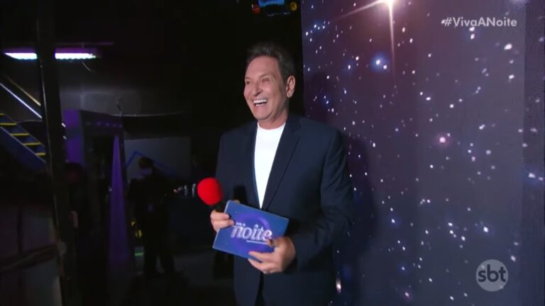 Luís Ricardo segura microfone e cartão do Viva a Noite nos bastidores do SBT com painel iluminado ao fundo.