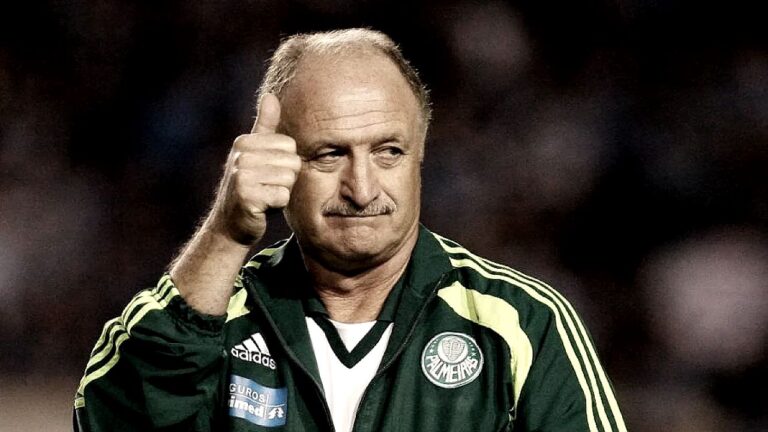 Homem mais velho, de bigode e cabelos grisalhos, veste agasalho esportivo verde e faz gesto de “joinha” com o polegar, em um ambiente que parece ser um estádio ou campo de futebol.