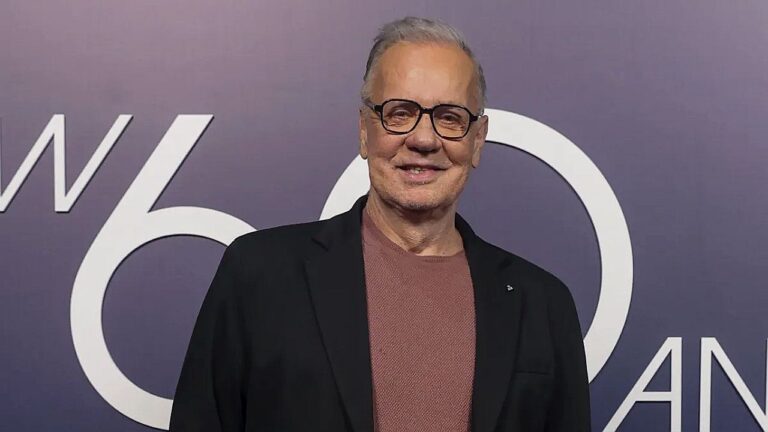 Homem de cabelos grisalhos e óculos sorri enquanto posa diante de painel de evento comemorativo, com número “60 anos” visível ao fundo