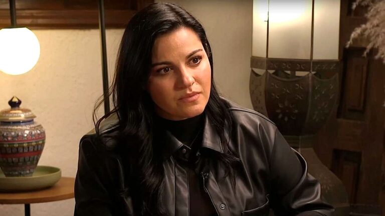 Mulher de cabelos escuros observa alguém com expressão séria durante conversa em ambiente interno iluminado, com luminária e objetos decorativos ao fundo