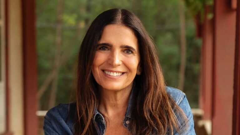 Atriz Malu Mader sorri interpretando Aurora no remake da novela Renascer, com cabelos longos escuros, usando jaqueta jeans em ambiente rural com árvores