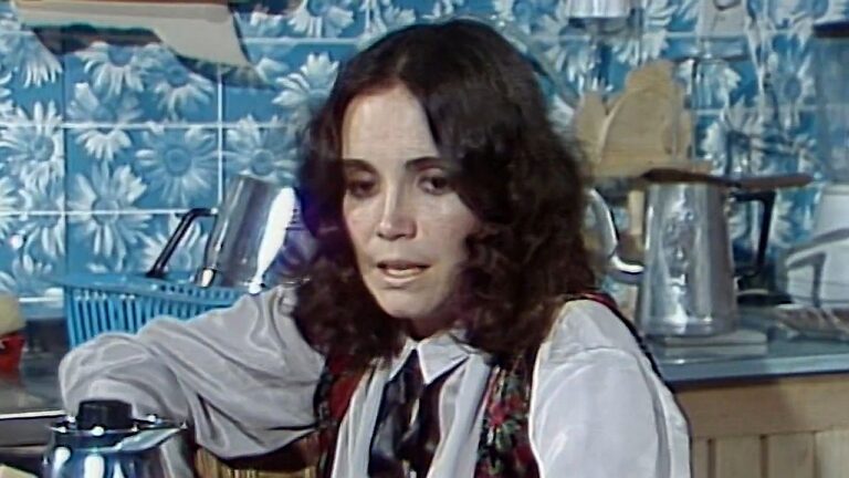 Mulher de cabelos escuros aparece preocupada em cena ambientada em cozinha com azulejos azuis e utensílios ao fundo