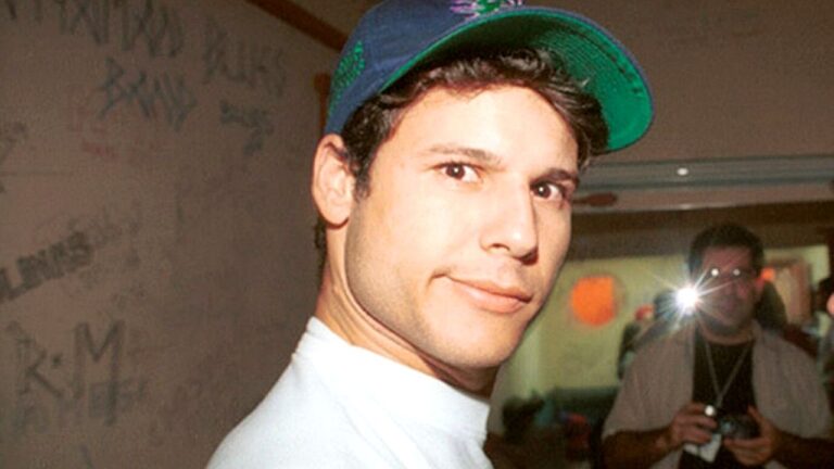 Homem jovem usa boné e olha para a câmera enquanto fotógrafo registra o momento com flash em ambiente interno de evento