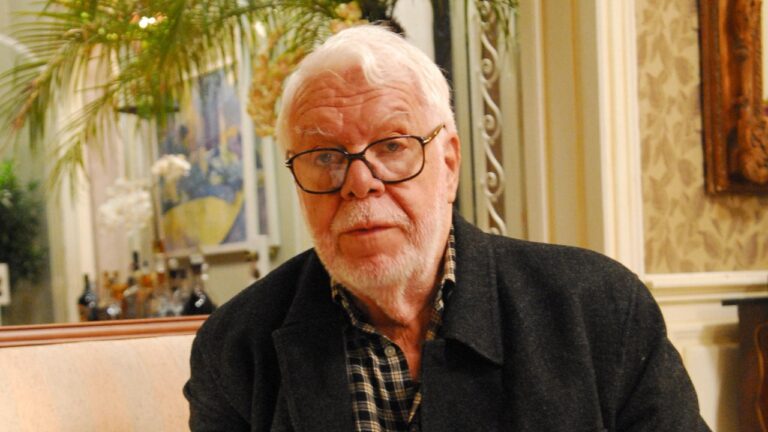 Autor Manoel Carlos, com cabelos e barba brancos, usa óculos de armação preta e casaco escuro sobre camisa xadrez, em ambiente interno decorado