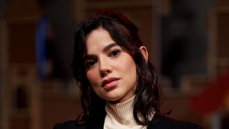 Mulher de cabelos castanhos escuros e expressão atenta aparece em close-up durante entrevista em estúdio, com iluminação suave e cenário interno desfocado ao fundo.