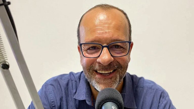 Um homem aparece em primeiro plano, sorrindo diretamente para a câmera. Ele usa óculos de armação escura, tem barba curta grisalha e cabelo com entradas. Está vestido com uma camisa azul e fala próximo a um microfone, sugerindo que está gravando ou participando de um podcast ou programa de áudio. Ao lado, é possível ver parte de um suporte de microfone. O fundo é simples e claro, sem muitos elementos, mantendo o foco nele.