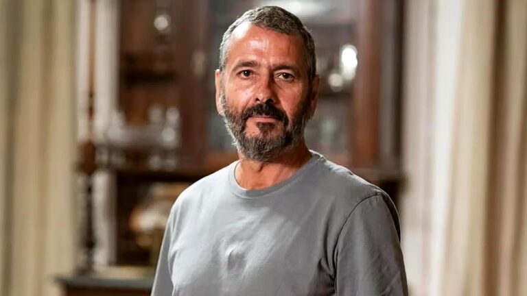 Um homem de meia-idade aparece em um ambiente interno, olhando diretamente para a câmera com uma expressão neutra e tranquila. Ele tem cabelos curtos grisalhos e barba também grisalha, bem aparada. Veste uma camiseta cinza simples. Ao fundo, há um móvel de madeira com portas de vidro, onde é possível ver objetos decorativos desfocados, sugerindo uma sala de estar ou jantar. A iluminação é suave e quente, destacando o rosto do homem.