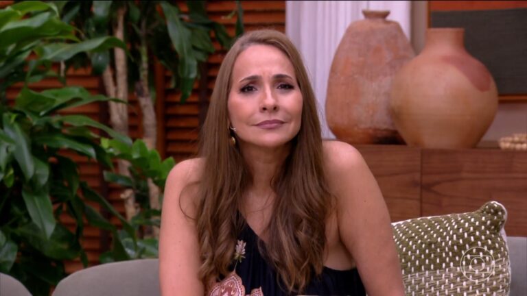 Maria Beltrão, com expressão irônica, fala no estúdio do É de Casa enquanto está sentada em um sofá. Ao fundo, há plantas e vasos decorativos, compondo um cenário de talk show; no canto da tela aparece o logotipo da TV Globo.