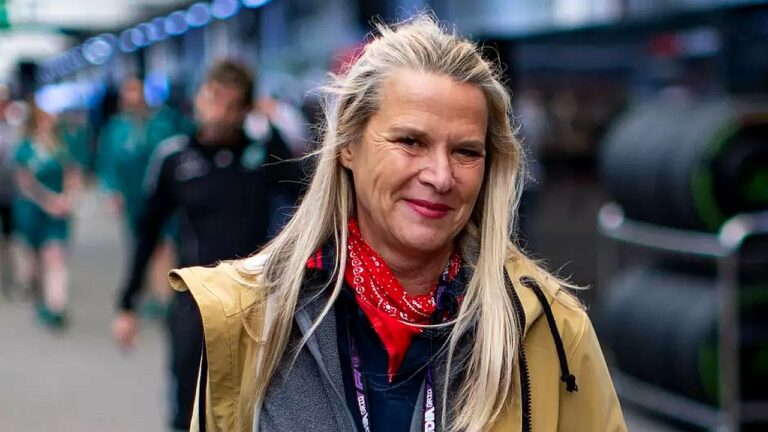 Texto Alternativo disse: Mulher de cabelo loiro e lenço vermelho caminha em paddock de automobilismo com boxes e equipe ao fundo