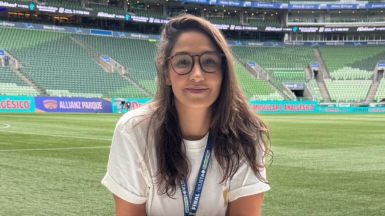 Mariana de Felippes sorri usando óculos de grau e camiseta branca no Allianz Parque, exibe crachá da Final Paulista 2024, gramado e arquibancadas ao fundo