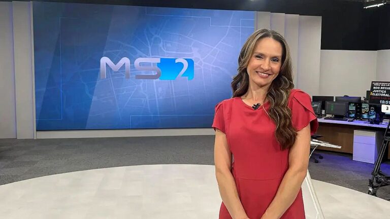 Mulher de vestido vermelho sorri em estúdio de telejornal com telão do MS2 ao fundo