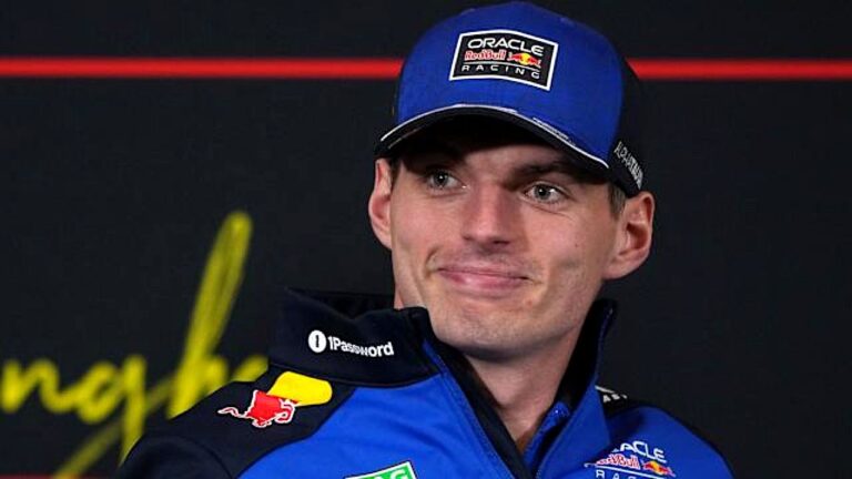Homem usando boné e uniforme de equipe de automobilismo, posando diante de um painel de patrocinadores em um ambiente de evento ou entrevista.