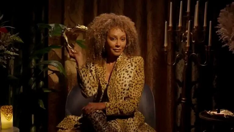 Mulher de cabelo cacheado veste roupa estampada de leopardo e segura taça dourada em ambiente com velas e decoração elegante ao fundo