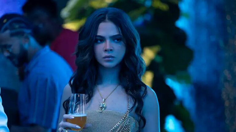 Mulher com vestido dourado segura taça e observa ao redor em ambiente noturno com luz azul