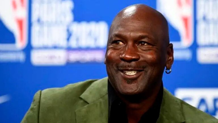 Homem careca sorri durante entrevista ou coletiva de imprensa, usando blazer verde e brinco, diante de painel azul com logotipos da NBA ao fundo.