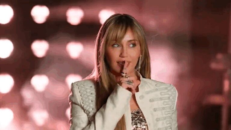 Miley Cyrus em um palco iluminado, com várias luzes desfocadas ao fundo criando um efeito brilhante. Ela usa um figurino elegante, incluindo um casaco claro com detalhes e uma roupa brilhante por baixo. Com expressão confiante e levemente provocativa, leva o dedo aos lábios em um gesto de silêncio. O cenário e a iluminação sugerem uma apresentação musical ou performance ao vivo.