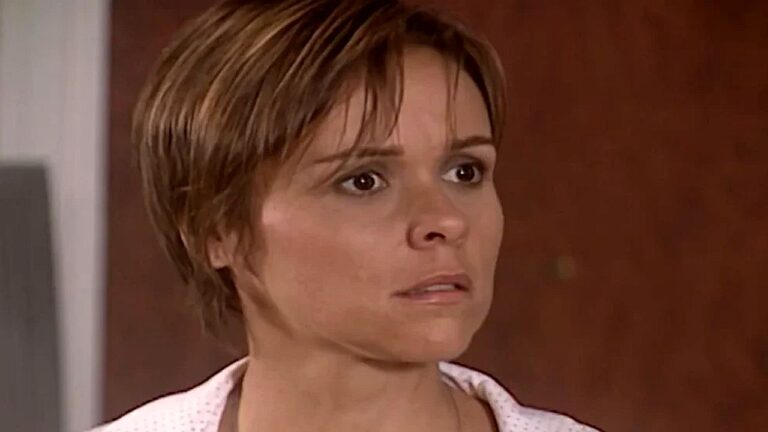 Mulher de cabelos curtos castanhos olha à frente com expressão séria e preocupada em cena interna com fundo neutro desfocado.