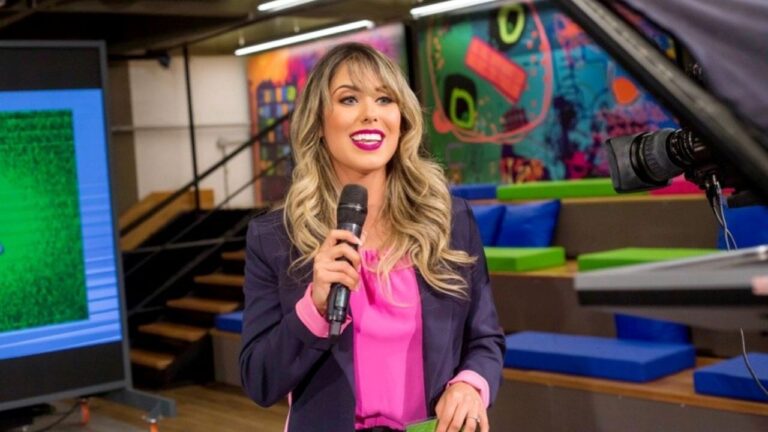 Myriã Pedron sorridente segura microfone preto, veste blazer azul marinho e blusa rosa choque em estúdio de TV com arquibancada colorida ao fundo
