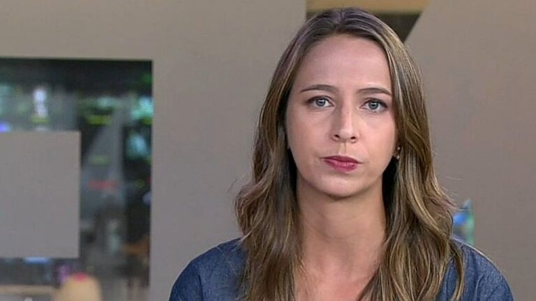 Uma mulher jovem de cabelo castanho claro, comprido e levemente ondulado está olhando diretamente para a câmera com expressão séria. Ela usa uma blusa em tom escuro. A imagem parece ser de uma cena interna, possivelmente em um ambiente doméstico ou escritório, com fundo desfocado que mostra paredes claras e uma abertura ou janela.