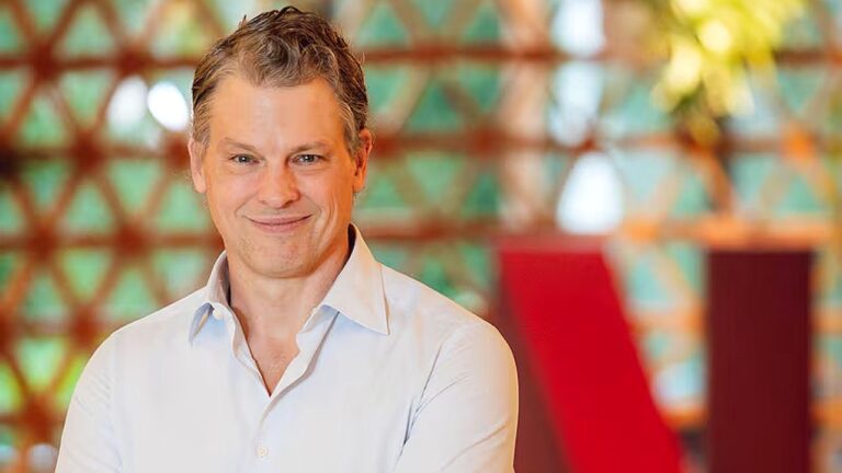 Homem de camisa clara sorri em retrato feito em ambiente interno com decoração moderna ao fundo