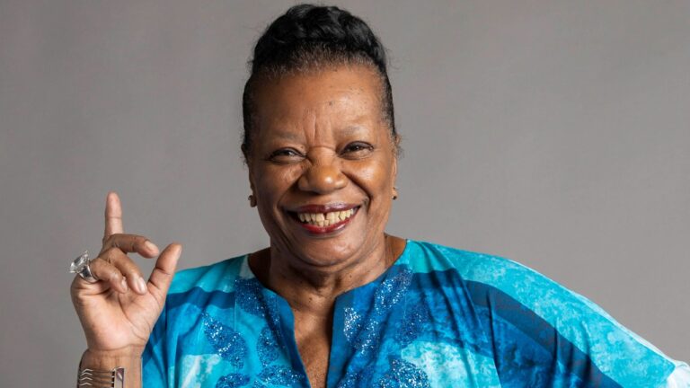 Atriz Neusa Borges sorri de bata azul estampada com detalhes brilhantes e aponta o dedo indicador para cima, usando anel de brilhante e fundo cinza