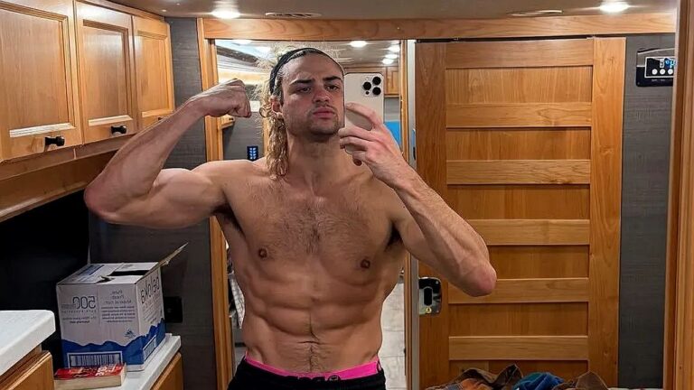 Homem sem camisa tira uma selfie no espelho enquanto flexiona o braço para mostrar o bíceps, em um ambiente interno com armários de madeira ao redor