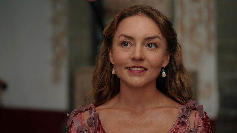 Mulher com vestido de época e brincos delicados sorri levemente em cena de novela de época em ambiente interno