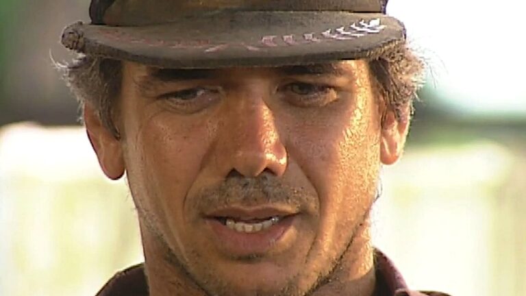 Homem usando chapéu aparece em close com expressão séria em cena ao ar livre sob luz do sol