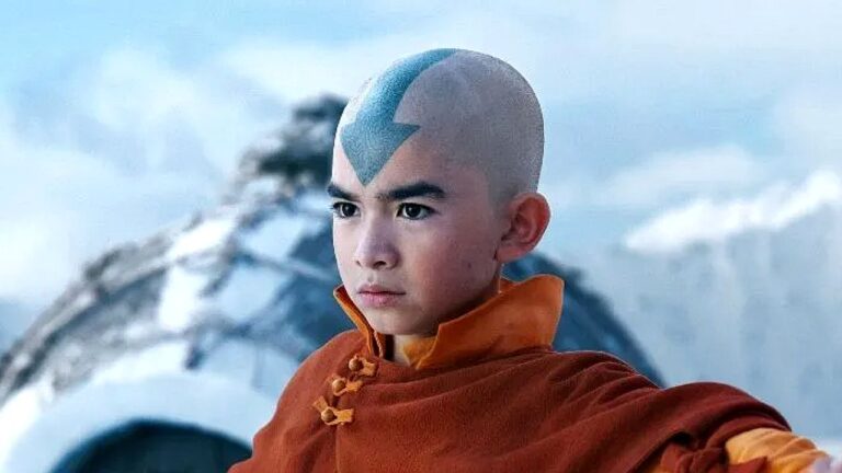 Personagem jovem com a cabeça raspada e uma seta azul pintada na testa, usando roupas de estilo monge, em um cenário frio com montanhas nevadas ao fundo