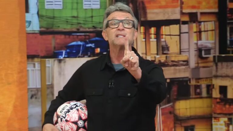 Craque Neto usa óculos e levanta o dedo indicador enquanto fala no estúdio de Os Donos da Bola, programa da Band, segurando uma bola de futebol debaixo do braço; ao fundo, há um painel com imagem de casas em uma comunidade.